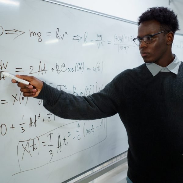 A serious math teacher in a classroom pointing at equations on a whiteboard.