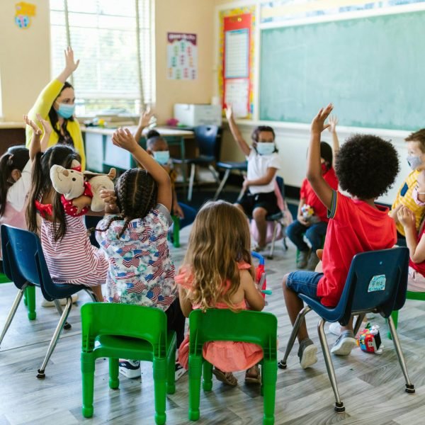 Diverse group of children in a classroom, raising hands, led by masked teacher. Bright and engaging learning environment.